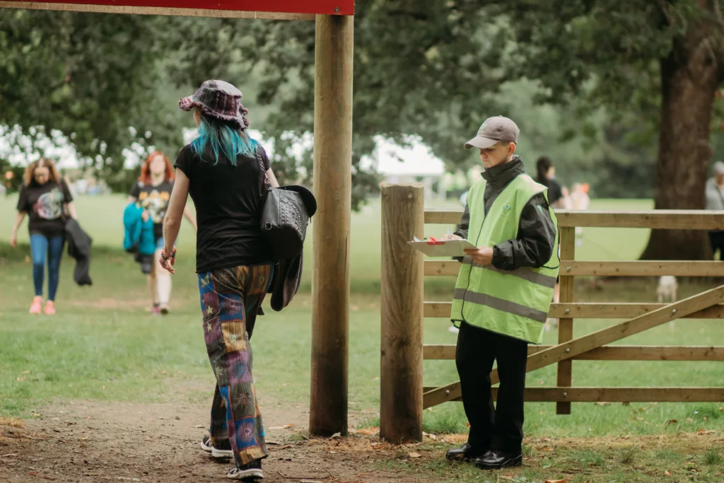 a steward in a high viz vest hold a clipboard while a festival goer, walks past the into the recovery festival
