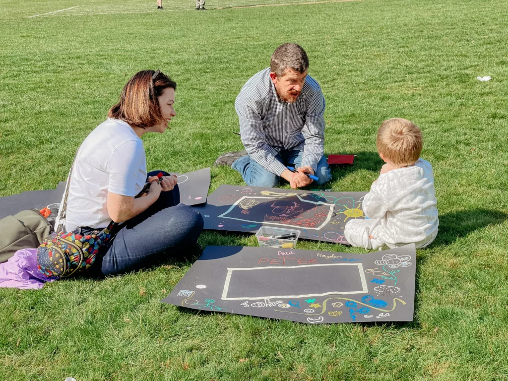 a young child talks with two people while sitting a grass they are helping him to draw something with chalk markers