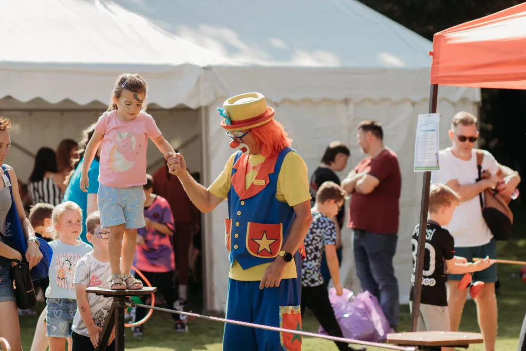 a clown helps a young girl cross a tightrope, while other children wait in line
