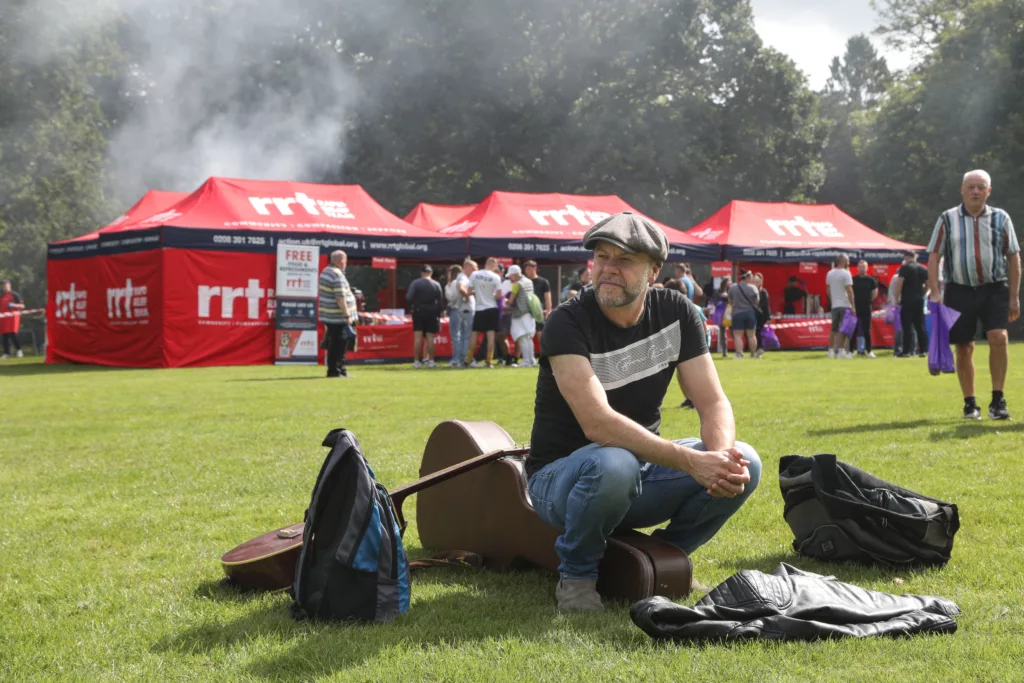 man sitting a his guitar case on the grass looking into distance while behind him food is cooked in tents